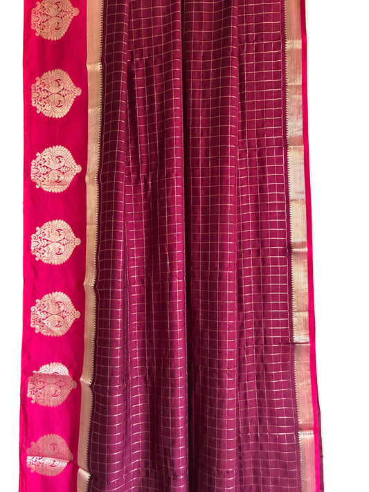A close-up of a traditional Indian saree fabric, showing a deep magenta body with a fine white grid pattern and a contrasting bright pink border adorned with gold zari motifs. aariyaa sarees for festive wear